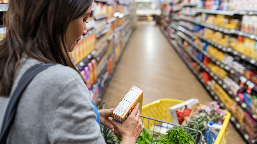 A woman in a grocery store aisle looks at the nutrition facts on an orange box as she leans on her yellow grocery cart. A woman in a grocery store aisle looks at the nutrition facts on an orange box as she leans on her yellow grocery cart.