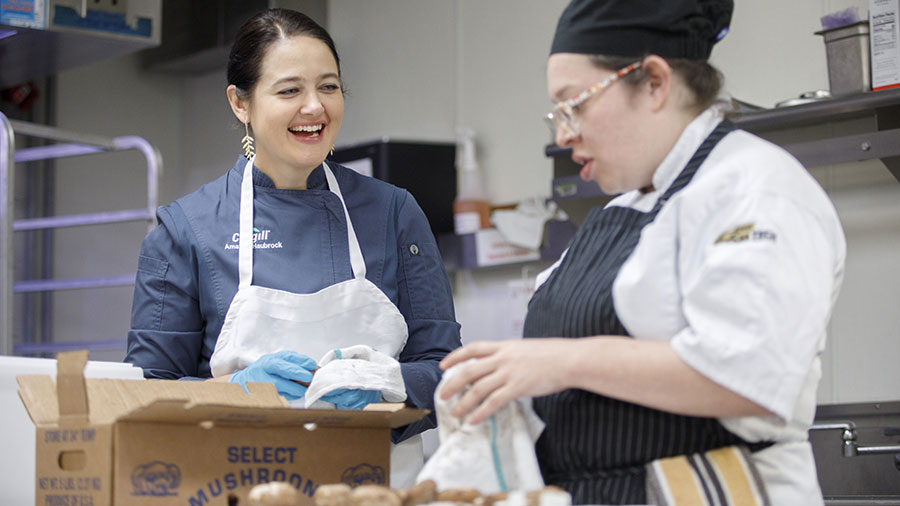 Two women chefs smile in a prep kitchen as they stand in front of a table with a pile of mushrooms wearing chef shirts and aprons. 