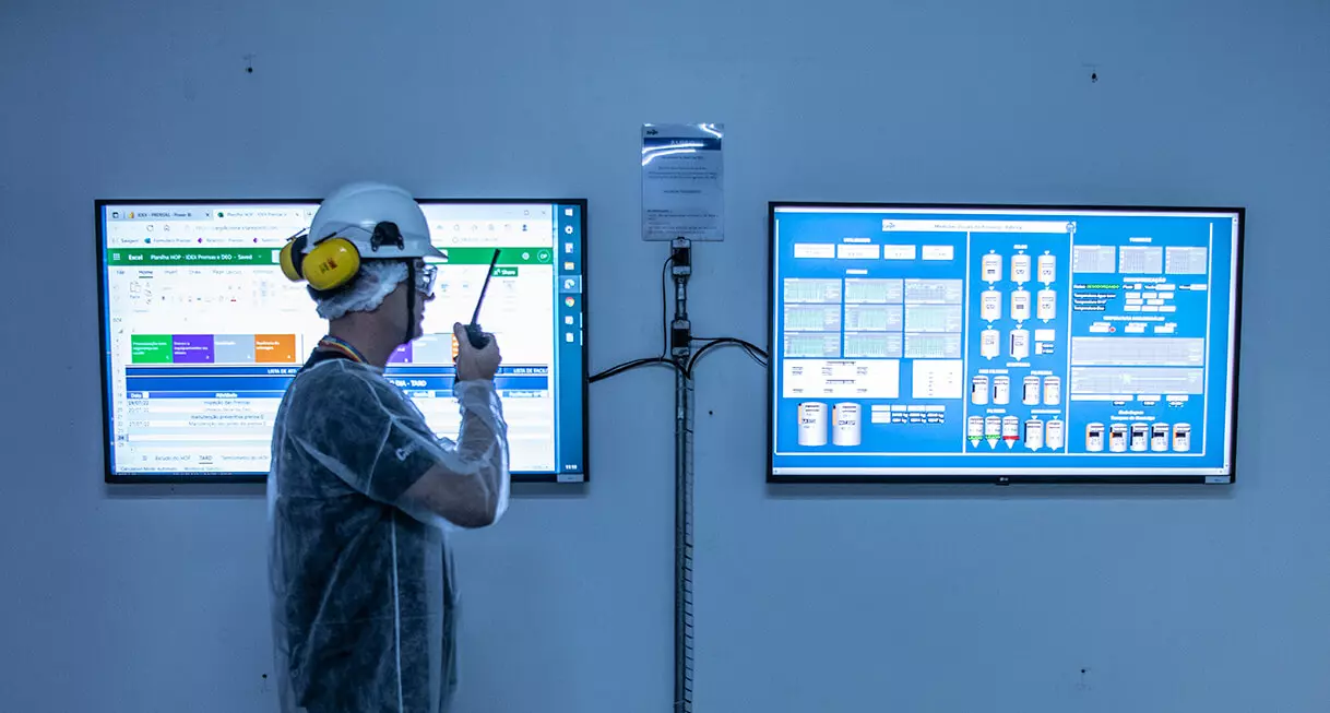 Photo of a Cargill employee speaking on a communication radio. He is standing, in profile, wearing a helmet and goggles, in front of two monitors with dashboards with production data