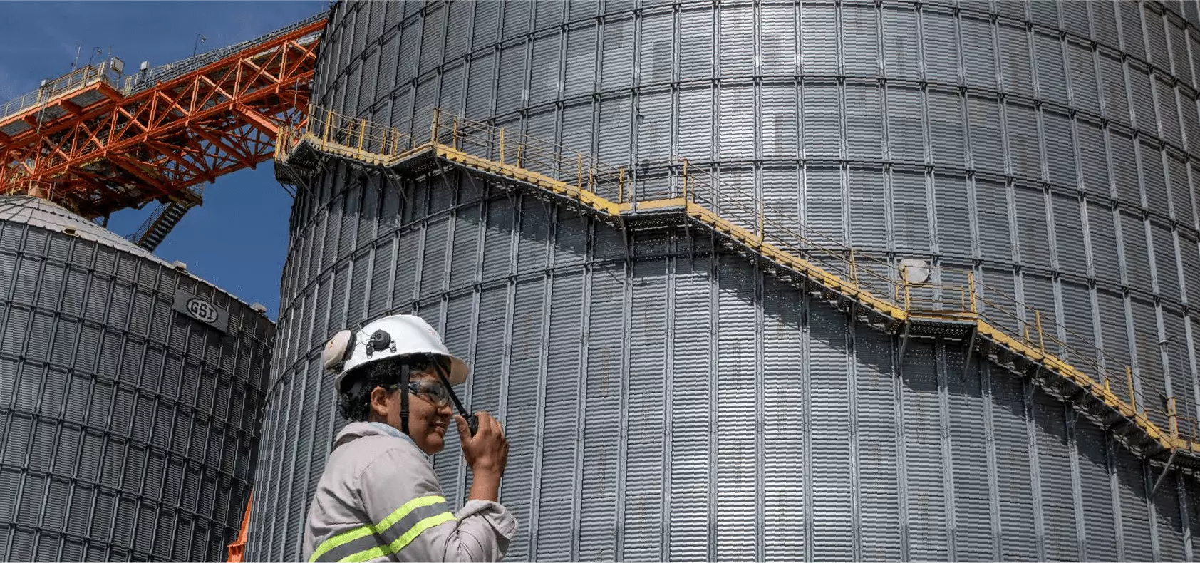 Foto de mulher, com capacete e óculos de proteção e rádio comunicador à mão, tendo ao fundo dois grandes silos de armazenamentos de grãos.